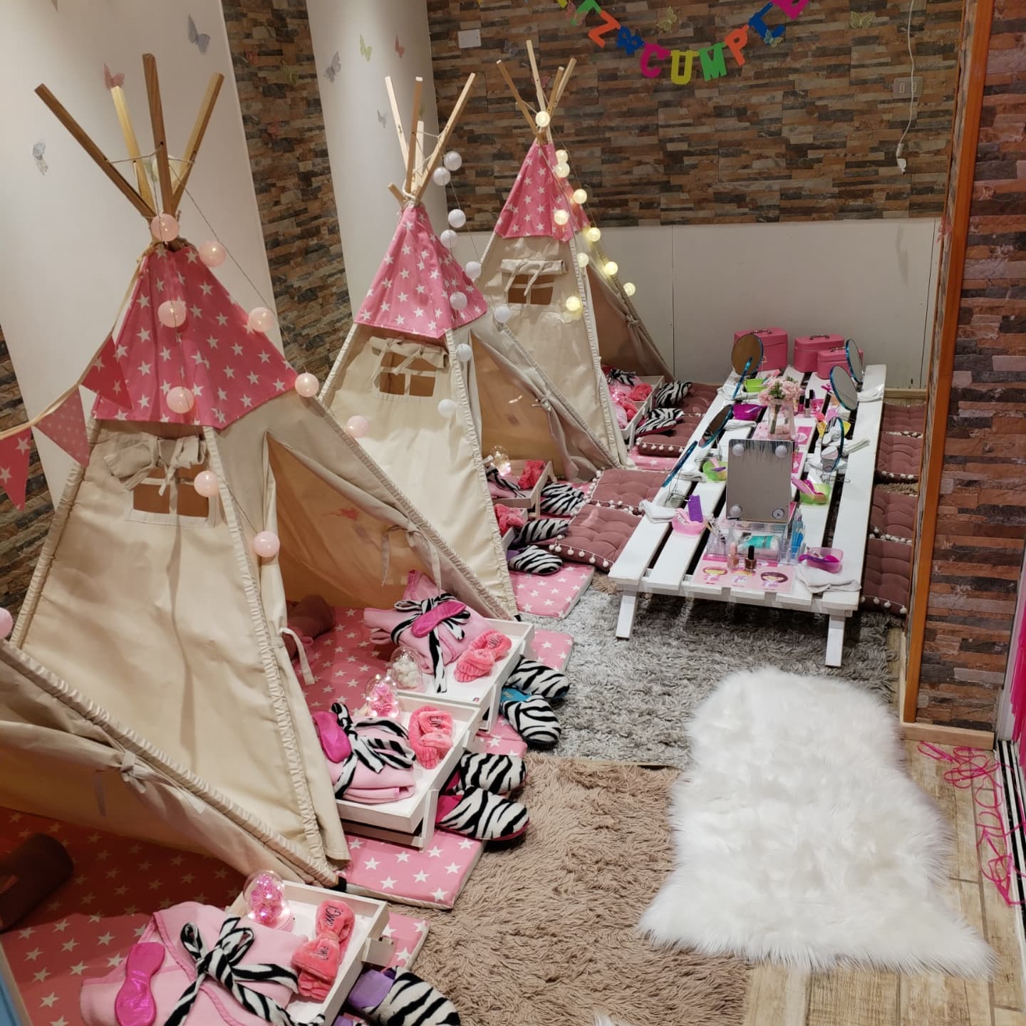 Carpas tipi decoradas con banderines y letrero de feliz cumpleaños, alfombra blanca y mesa de pallets con kits de spa